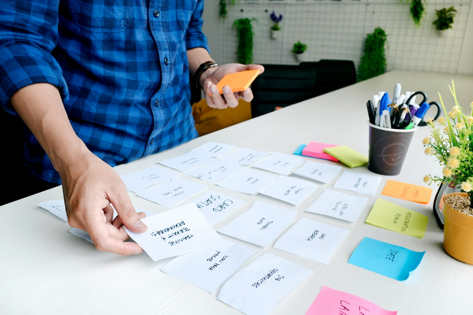 A person in a blue plaid shirt organizes handwritten notes on a workspace, focusing on planning or brainstorming activities. Colorful sticky notes and office supplies are visible.
